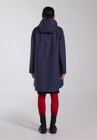 Námořnicky modrý nepromokavý trenčkot s kapucí, dlouhými rukávy a volným střihem, doplněný červenými punčocháči a černými kotníkovými botami.