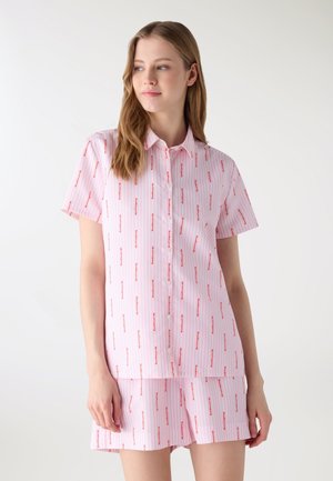 Giovane donna che indossa una camicia a maniche corte a righe rosa e bianche con bottoni e pantaloncini abbinati con piccolo motivo di testo rosso, in piedi davanti a uno sfondo bianco.