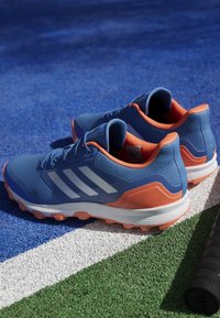 Blå sportskor med orange detaljer, texturerad mesh-övre del, tre vita ränder och en robust sula för grepp. Positionerade på en texturerad yta.