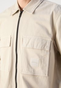 Jasno beżowa kurtka z zamkiem błyskawicznym z przodu, dwoma kieszeniami na piersi oraz kwadratową naszywką z logo. Wykonana z gładkiego materiału z klasycznym kołnierzem i przeszyciami.