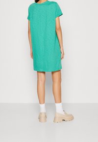 T-Shirt-Kleid mit kurzen Ärmeln in Teal, entspannter Schnitt, runder Ausschnitt und geradem Saum. Getragen mit beigen Plateau-Schuhen und weißen Socken.