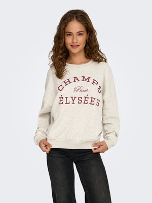 Ung kvinde med krøllet hår iført en cremefarvet sweatshirt med "Champs Paris Élysées" i rødt, kombineret med sorte jeans, stående mod en hvid baggrund.