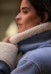 Giubbotto di jeans con colletto e polsini in shearling crema e soffice. Presenta una vista ravvicinata che evidenzia la texture e il contrasto dei colori.