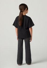 Otrok z dolgimi rjavimi lasmi v košarici, oblečen v črno majico in temno sivo jeans, stoji obrnjena proti enostavni svetlo sivi ozadju.