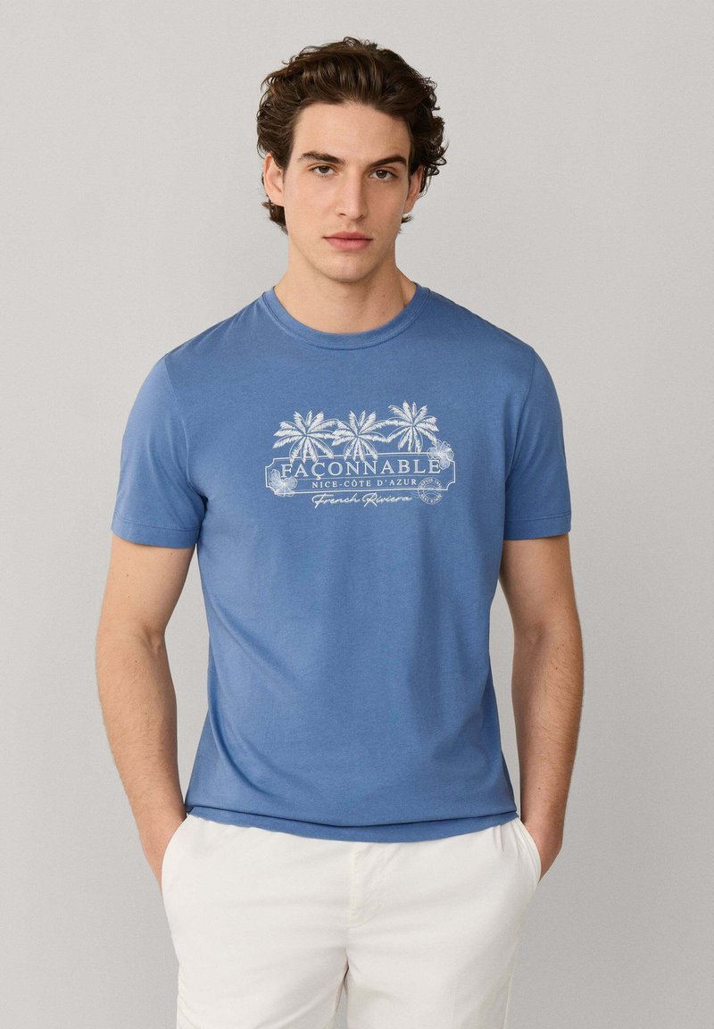 Mann trägt ein blaues Façonnable-T-Shirt mit weißem Palmenmotiv und weiße Hose, steht mit Händen in den Taschen vor einem einfarbigen Hintergrund.