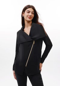 Juoda striukė su dideliu, drapiruotu apykakle, asimetrišku priekinio užtrauktuko uždarymu ir dviem šoniniais kišenėmis; pagaminta iš lygios medžiagos su elegantiška tekstūra.