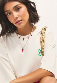 Cream cotton t-shirt features short sleeves and a green checkerboard design. Necklaces with red chili pepper pendants, gold chain accents.