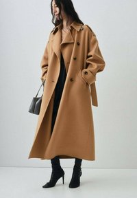 Long camel coat with a wide collar and double-breasted design. Soft fabric, two large side pockets, and a tied waist detail. Black high heel boots.