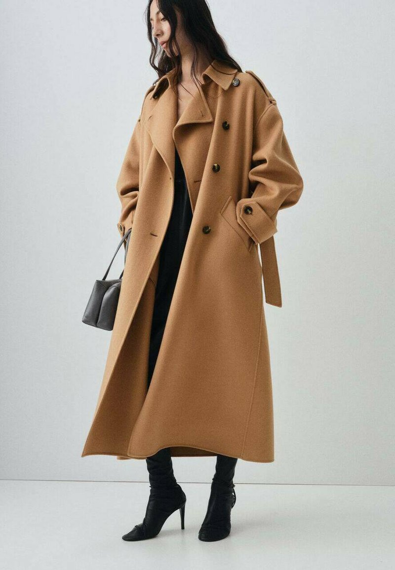 Long camel coat with a wide collar and double-breasted design. Soft fabric, two large side pockets, and a tied waist detail. Black high heel boots.