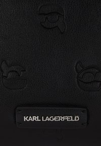 Crna kožna torba s utisnutim logotipima Karl Lagerfeld i srebrnom pločicom s logotipom. Tekstura površine s minimalističkim dizajnerskim detaljima.