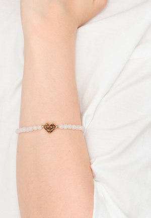 Wrist with white beaded bracelet featuring a metallic geometric heart charm worn over a white fabric background.