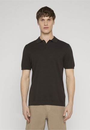 Ung mand, der vender fremad, iført en sort polo med korte ærmer og beige bukser mod en ensfarvet lys baggrund.