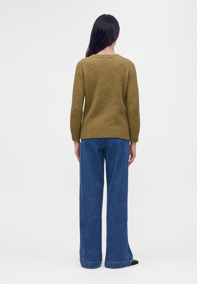 Femme debout, de dos, portant un pull vert olive et un jean bleu à jambes larges sur un fond clair uni.