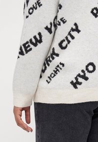 Vaatteena on vaalea neulepaita, jossa on mustia kaupunkien nimiä, kuten "New York" ja "Kyoto". Ribbaatut hihat ja helma; tekstuuri, rento malli.