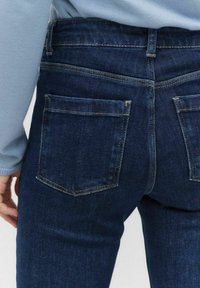 Tummansiniset farkut, joissa on kapea malli, kahdella takataskulla ja kontrastivärillä ommellut saumat. Kangas näyttää sileältä, jossa on minimaalinen rakenne.