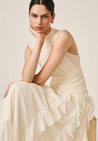 Joven mujer con cabello oscuro con un vestido de color marfil sin mangas y cuello alto, con cintura fruncida y falda con volantes, apoyando la barbilla en la mano mientras está sentada.