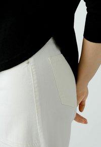 Weiße Jeans mit einer Gesäßtasche und sichtbaren Nahtdetails, ergänzt durch ein schwarzes Oberteil, das teilweise den Taillenbund verdeckt.