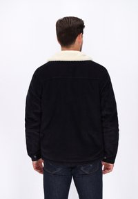 Schwarze Cordjacke mit einem weißen Kunstfellkragen, ausgestattet mit einem Knopfverschluss und zwei Taschen. Die Ansicht zeigt die Rückseite des Kleidungsstücks.