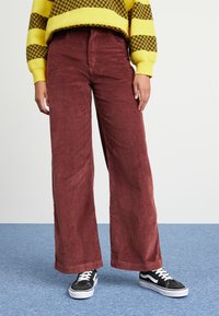 Persoon in een wijde maroon corduroy broek, zwart-witte sneakers en een gele trui met een bruin houndstooth- en streep patroon.