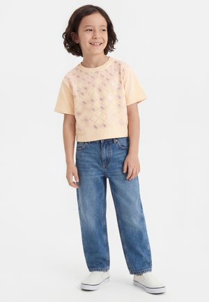 Enfant aux cheveux mi-longs portant un haut à manches courtes à motifs pêche, un jean bleu et des chaussures blanches sans lacets, debout et souriant.