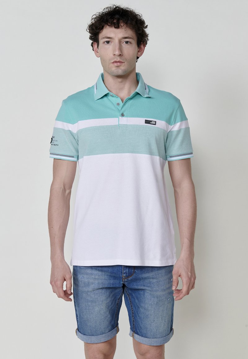 Világos teal színű polo póló vízszintes fehér csíkokkal, rövid ujjú és galléros. A mellkasán egy kis logó folt található. Farmer sorttal viselve.