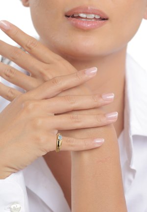 Kvinde med lyserødt polerede negle iført en guldring med en lille hjerteformet ædelsten på fingeren, hænderne rører hendes håndled.