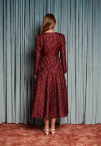 Femeie stând cu fața spre draperii albastre, purtând o rochie midi bordo cu flori și mâneci lungi și pantofi cu toc albi pe o mochetă maro.