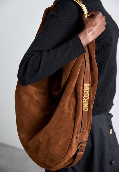 Sac hobo en daim brun avec une texture lisse, quincaillerie dorée et logo "MOSCHINO" en lettres majuscules, présenté sur un haut en tissu côtelé noir.