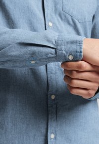 Lichtblauw overhemd met knopen en een zachte textuur, voorzien van een borstzak en manchetten met knopen. De knopen zijn wit en rond.