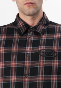 Camicia a quadri nei toni del nero, rosso e grigio, con un design bottone, colletto a punta e una toppa logo nera testurizzata sul petto.