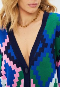 Knit cardigan featuring a deep V-neck, blue, green, pink, and black geometric patterns, with a central button detail. Gold chain necklace included.