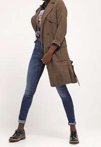 Brązowy zamszowy płaszcz trench z dwiema dużymi kieszeniami, w połączeniu z ciemnoniebieskimi dopasowanymi dżinsami i błyszczącymi czarnymi sznurowanymi butami.