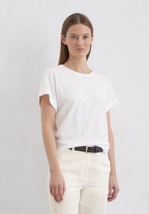 Camiseta blanca de manga corta con puños enrollados, hecha de una tela ligera, combinada con pantalones de color claro y un cinturón negro con herrajes dorados.