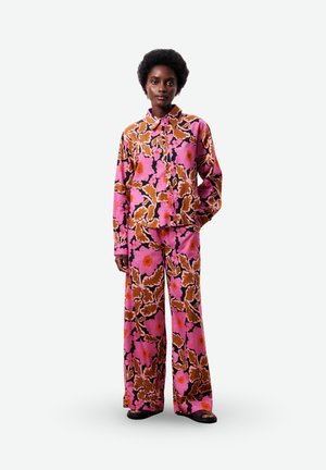 Femme portant une chemise à manches longues et un pantalon large à imprimé floral assorti rose, marron et orange, debout avec une main dans la poche.