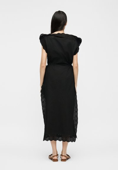 Γυναίκα με μακριά σκούρα μαλλιά που φοράει μαύρο φόρεμα μέχρι τη μέση της γάμπας με διακόσμηση από δαντέλα με μικρές οπές, όρθια ξυπόλυτη σε λευκό απλό φόντο.