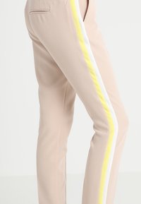Beige trousers with a slim fit, featuring a yellow and white side stripe. Made from a smooth fabric, they have a tailored design and pocket details.