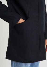 Persona che indossa un cappotto di lana nero con una mano nella tasca laterale, abbinato a jeans celesti, su uno sfondo semplice.