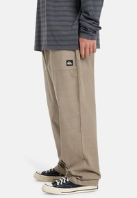 Beige, loose-fitting pants with a side pocket and a black logo patch. Paired with black sneakers featuring white rubber soles.