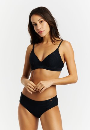 Žena stoji na bijeloj pozadini noseći crni bikini s diskretnom teksturom, ruke joj su opušteno spojene sprijeda.