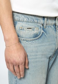 Light blue denim jeans with a faded texture, featuring a small logo tag, and a silver chain bracelet on a hand resting near the pocket.