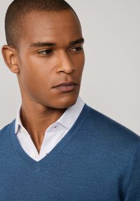 Maglione blu a v con tessuto fine a maglia, indossato sopra una camicia bianca con strisce azzurre e colletto a bottoni.
