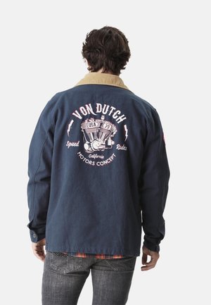 Homme portant une veste bleu marine avec un col beige, dessin graphique de moteur de moto et texte « Von Dutch, Speed Rules, California Motors Concept » dans le dos.