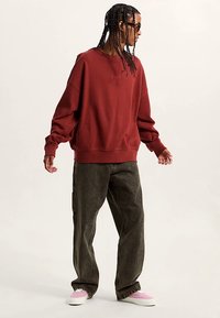 Vinytrød sweatshirt med oversized pasform og ribstrikkede manchetter, kombineret med mørkegrønne løse bukser og pink slip-on sko, vist fra siden.