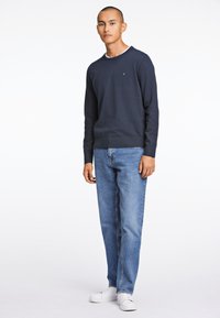 Pull en maille bleu marine avec col rond, associé à un jean droit bleu clair et des baskets blanches. Le pull présente un petit logo.