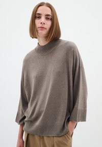 Per didelis taupe spalvos megztinis su aukštu kaklu, laisvu reguliavimu ir raukšlėtais rankogaliais. Minkšta tekstūra ir minimalus dizainas, derinamas su bežinėmis kelnėmis.