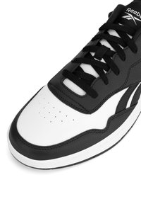Czarno-białe buty sportowe z gładką skórzaną cholewką, kontrastowymi teksturowanymi akcentami, perforowanym czubkiem i czarnymi sznurówkami. Biała gumowa podeszwa.