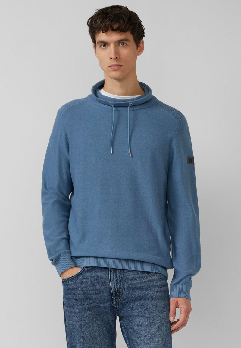 Jasnoniebieski sweter z golfem, z wysokim, złożonym kołnierzem, sznurkami regulacyjnymi, prążkowanym dołem i mankietami oraz naszywką na lewym rękawie.