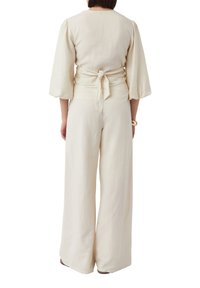 Jumpsuit w kolorze kremowym, wykonany z lnu, z szerokimi nogawkami, ściągniętą talią związaną z tyłu oraz rękawami o długości trzy czwarte, z gładką teksturą.