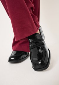 Scarpe in pelle verniciata nera con punta rotonda, dotate di due cinturini in velcro, esposte accanto a pantaloni di velluto a coste color bordeaux.