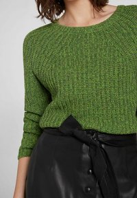 Frau in einem strukturierten grünen Strickpullover mit rundem Ausschnitt, der in einen schwarzen Lederrock gesteckt ist, der mit einem großen Schleifen-Gürtel gebunden ist.